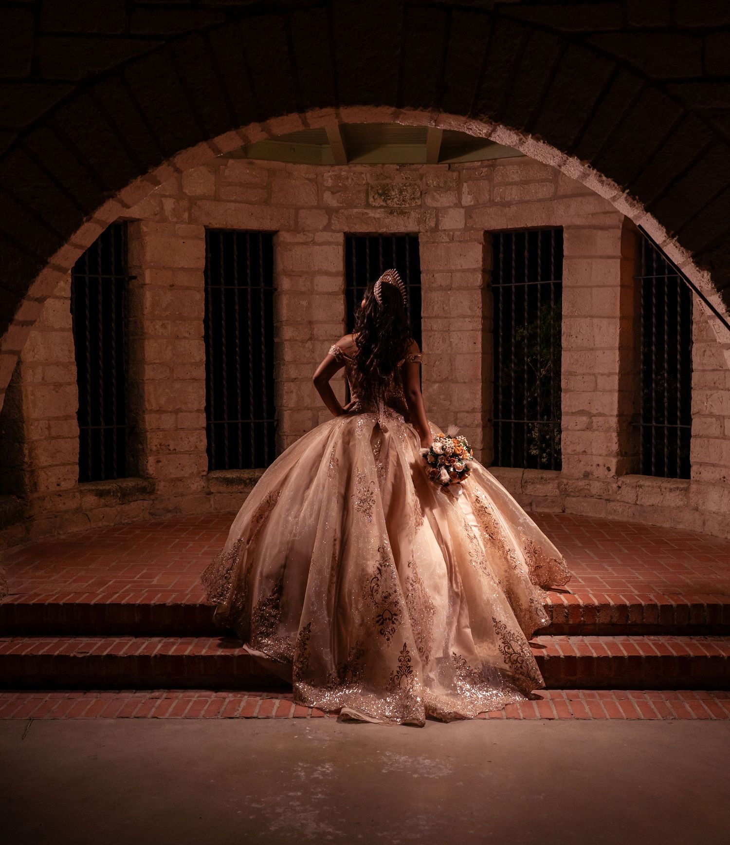 Bride in elegant ballgown stands beneath stone arch, holding bouquet, facing ornate windows in dramatic lighting.