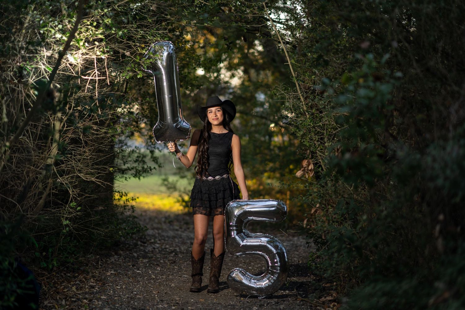 Young woman in black dress and cowboy hat holding silver number 15 balloons on a forest path.