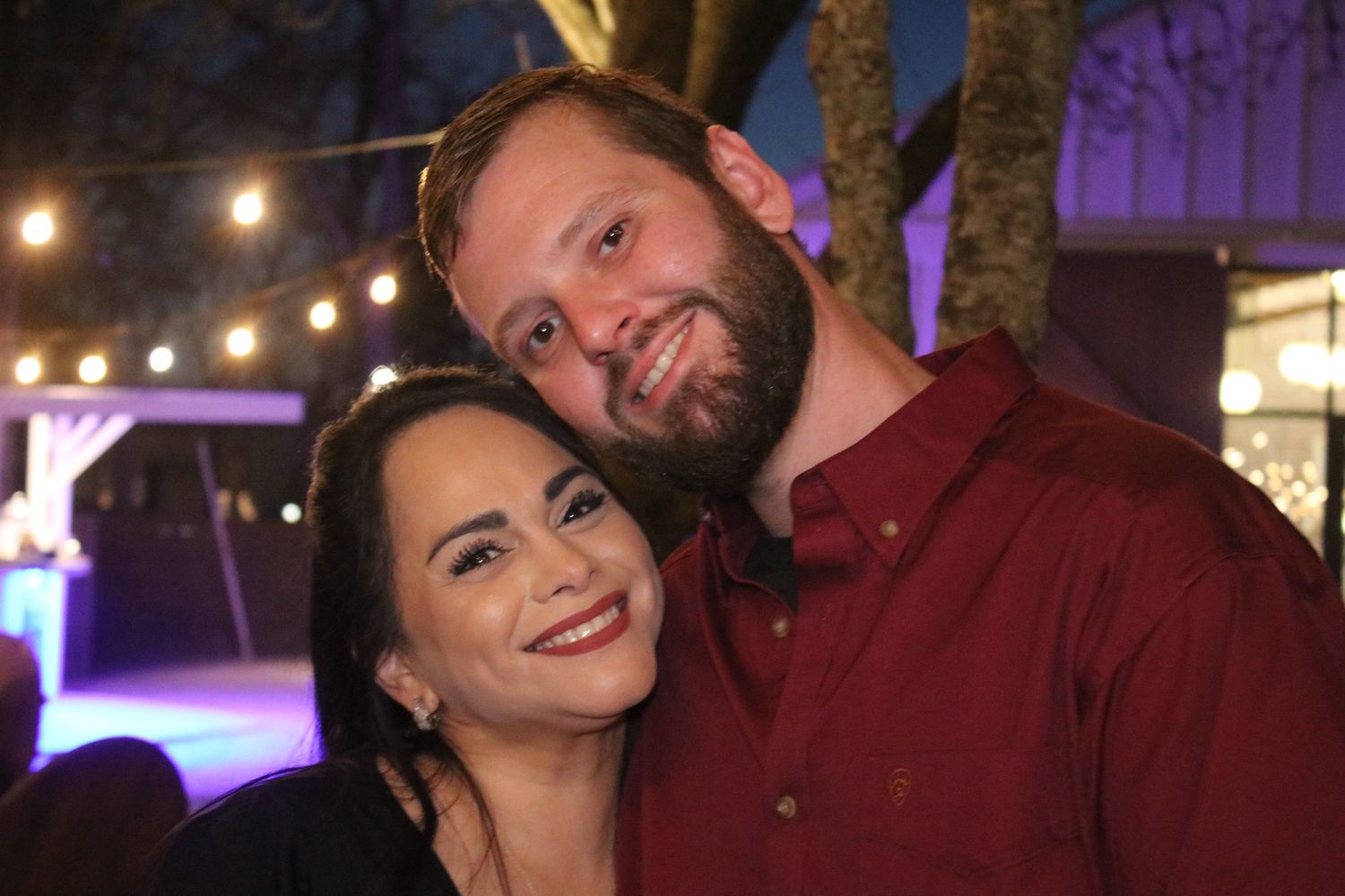 A couple smiles together at a nighttime event with purple lighting and string lights in background.