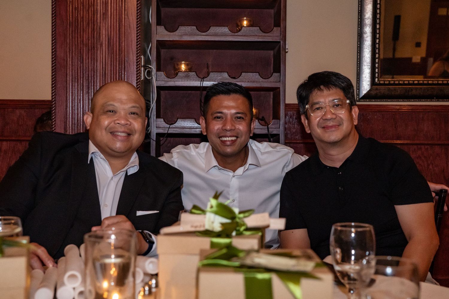 Three smiling men pose together at a restaurant dinner table with wine glasses and elegant decor.