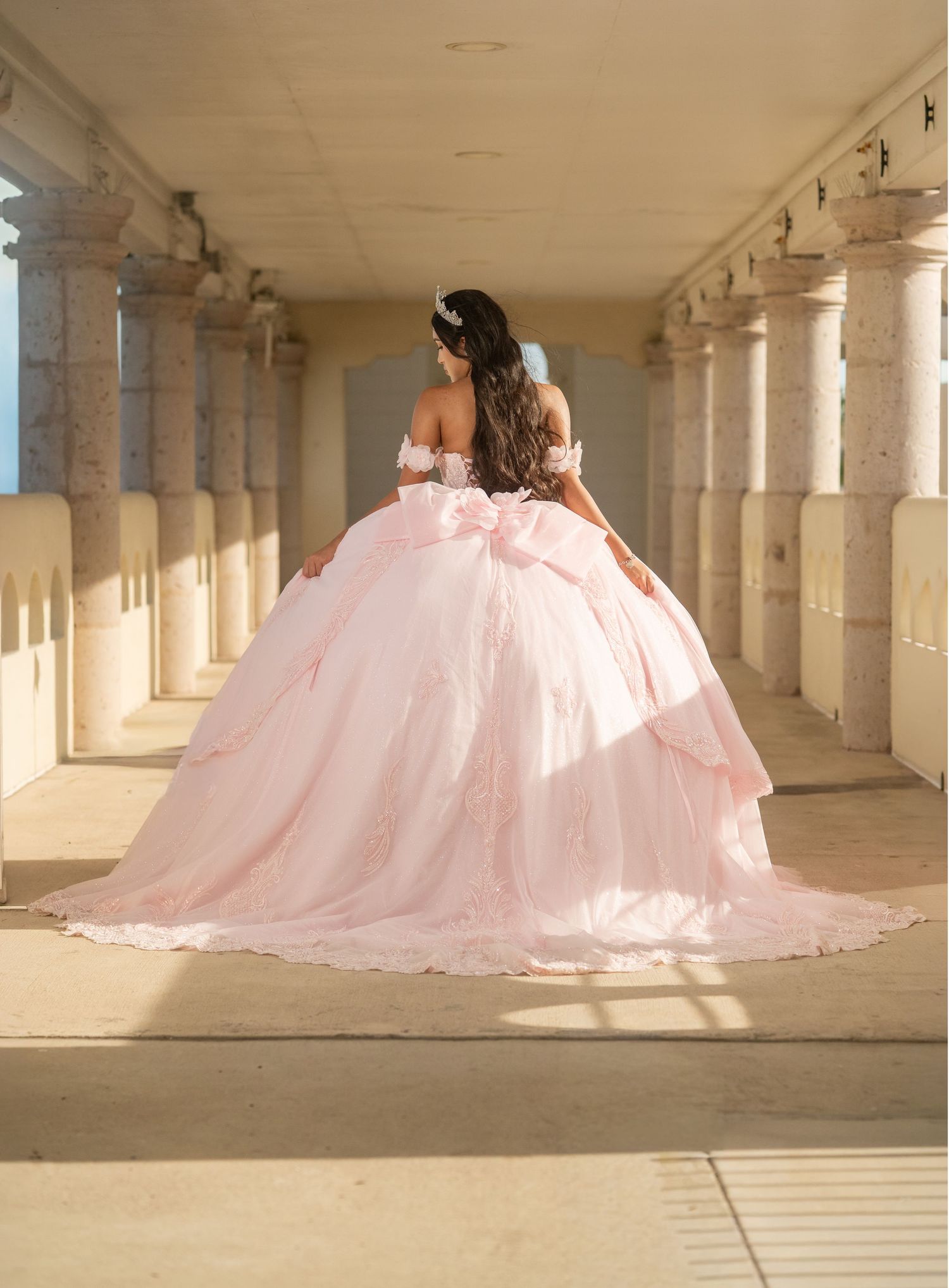 Young woman in a stunning blush pink off-shoulder ball gown with large bow, posing in an elegant columned hallway.
