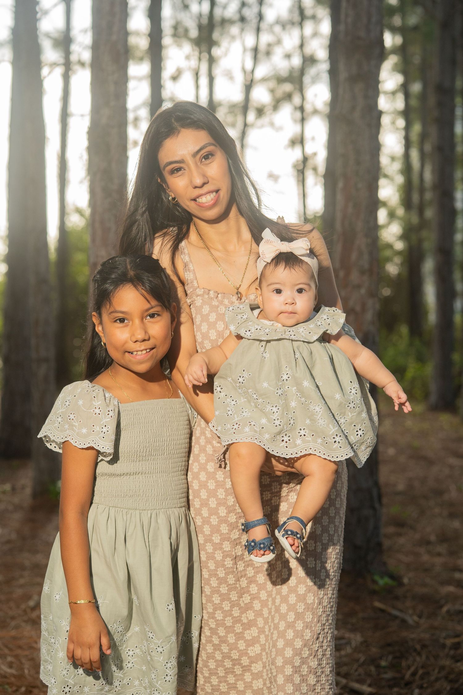 A mother holds her baby girl while standing with her older daughter in a sunlit pine forest, all wearing coordinating dresses.