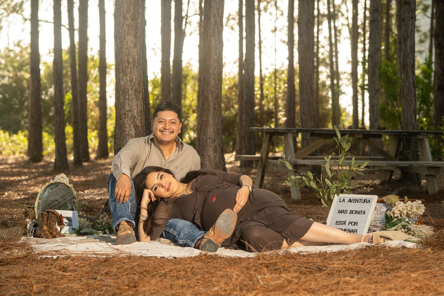 Two women relaxing on a blanket in a sunlit pine forest, one lying down smiling, the other sitting beside her.