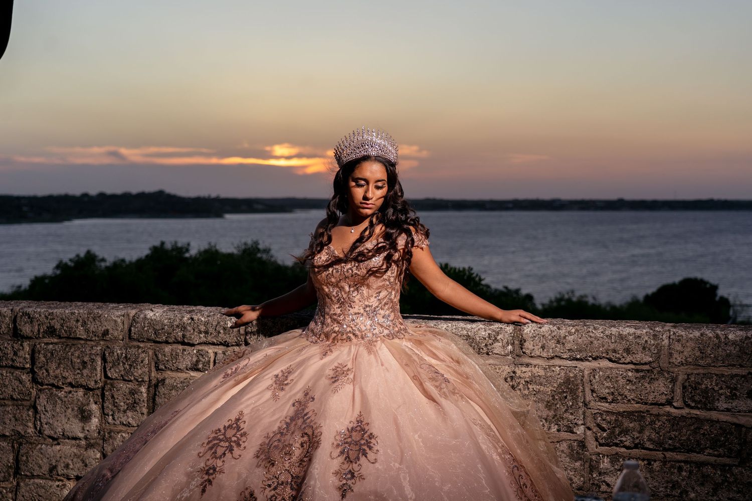 Person wearing a blush pink quinceañera ball gown with beaded bodice poses at sunset by stone wall overlooking water.