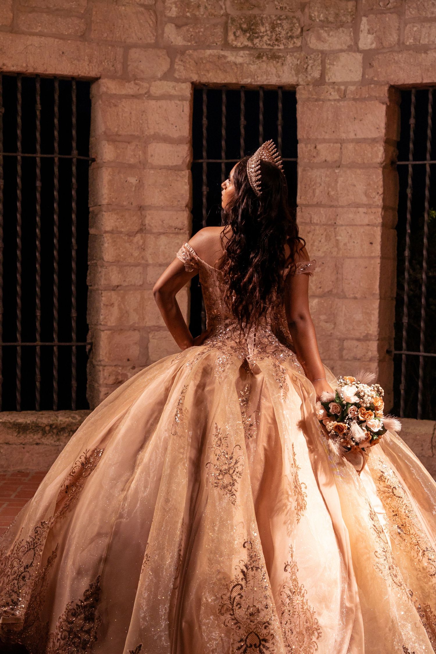 Back view of an ornate champagne-colored quinceanera gown with intricate beading against stone fortress wall.