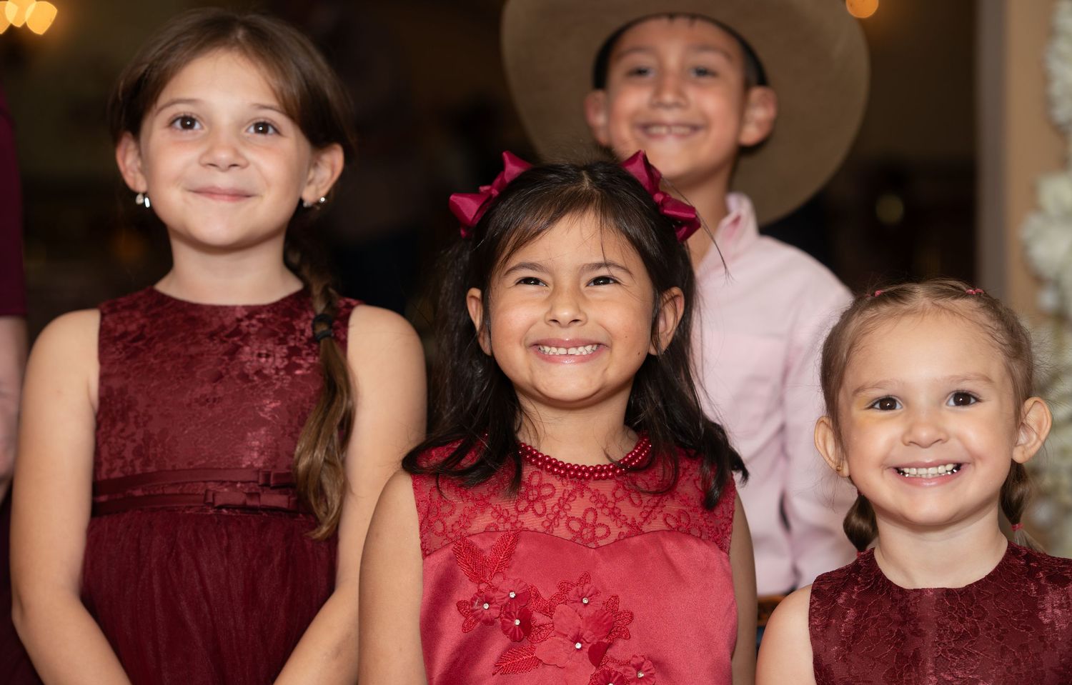 Four young children in red dresses and formal wear smiling at a quinceañera party.