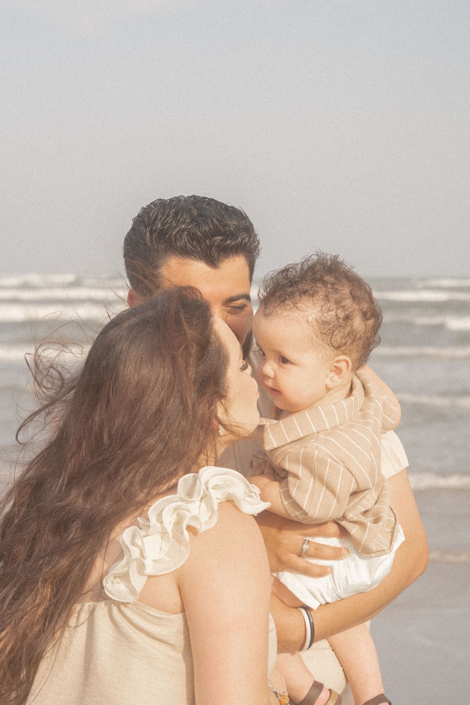 Parents sharing tender moment kissing their baby at the beach on a sunny day.