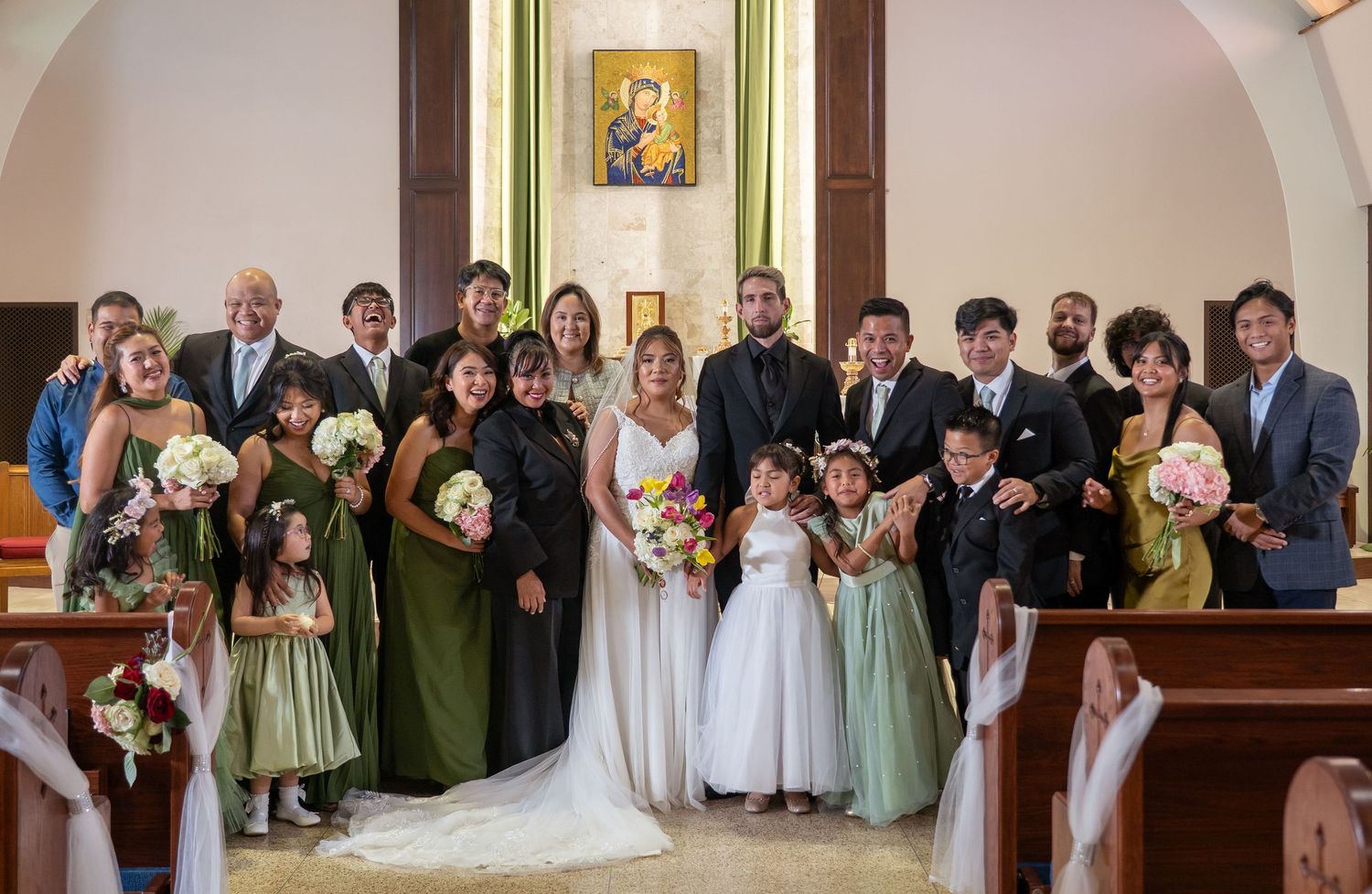 Wedding party poses in a church with bride and groom centered, bridesmaids in green dresses, and flower girls in white.
