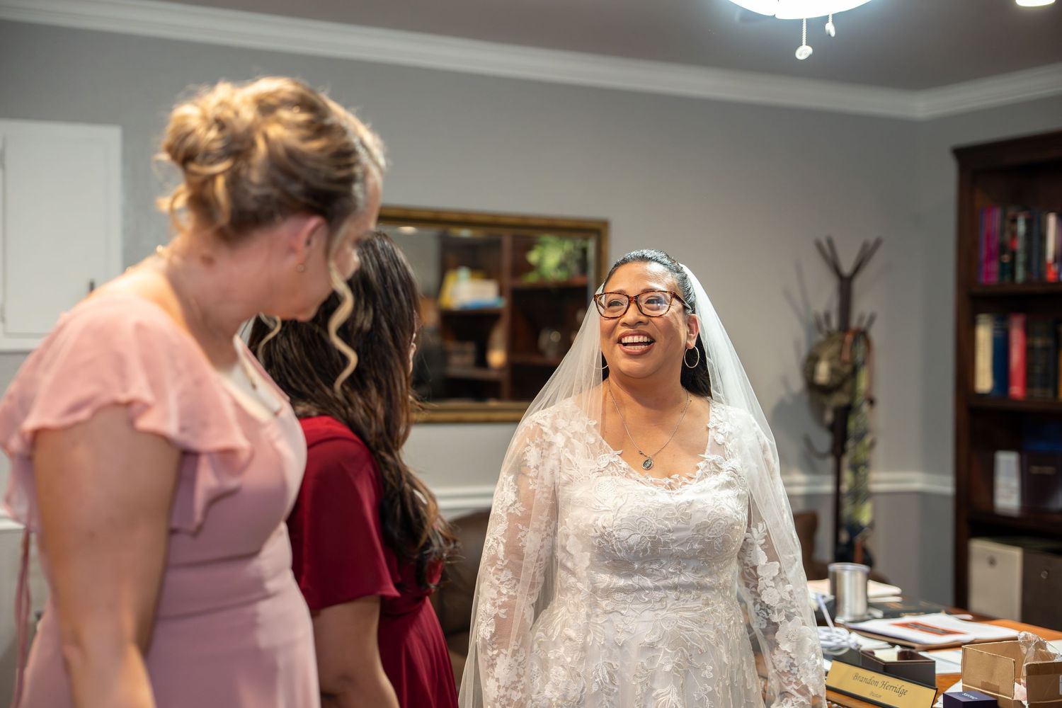 Bride in white lace wedding gown and veil laughing joyfully with bridesmaids in pink and burgundy dresses.