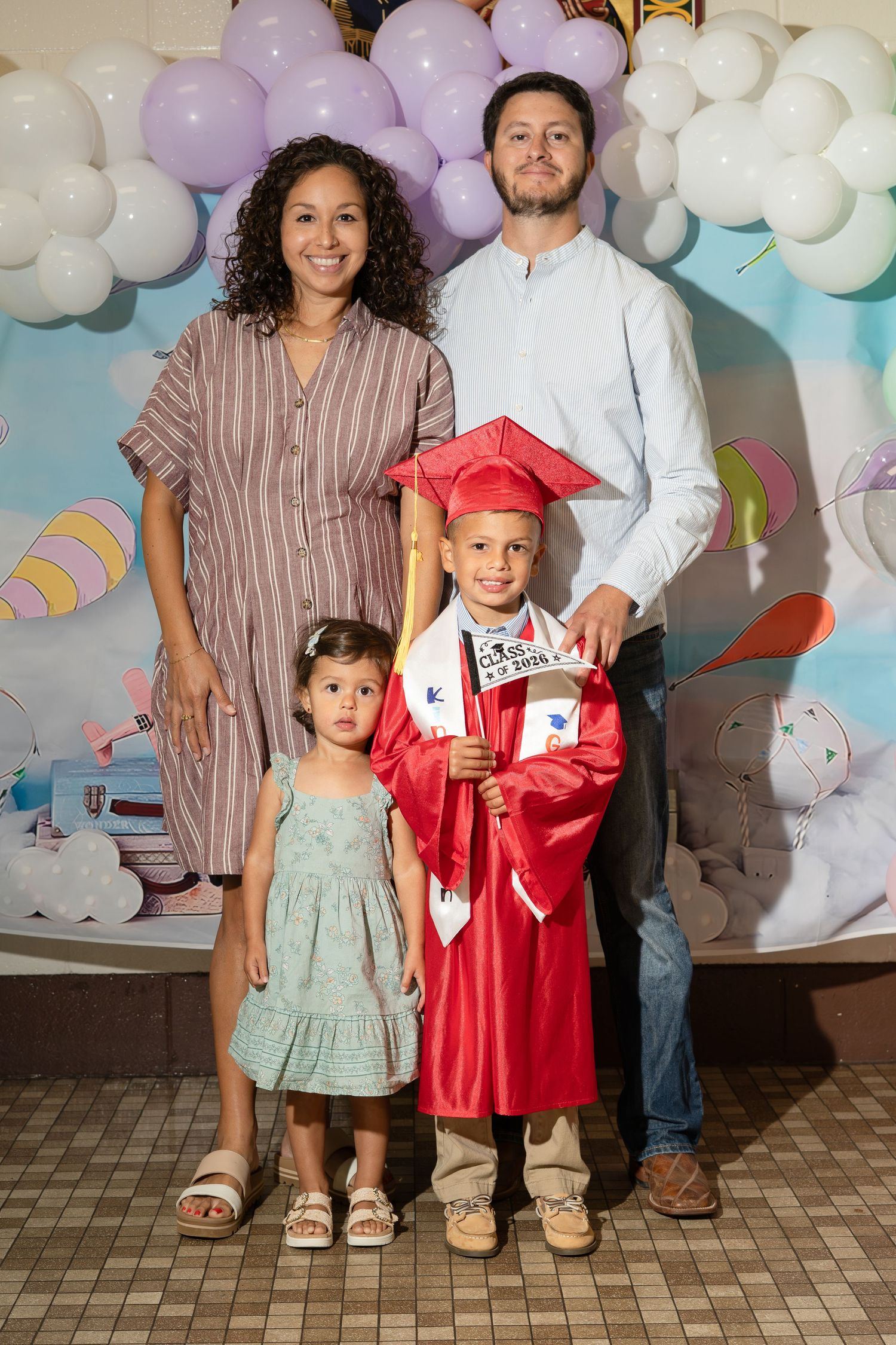 Family of four celebrates young boys kindergarten graduation in red cap and gown with balloon backdrop.