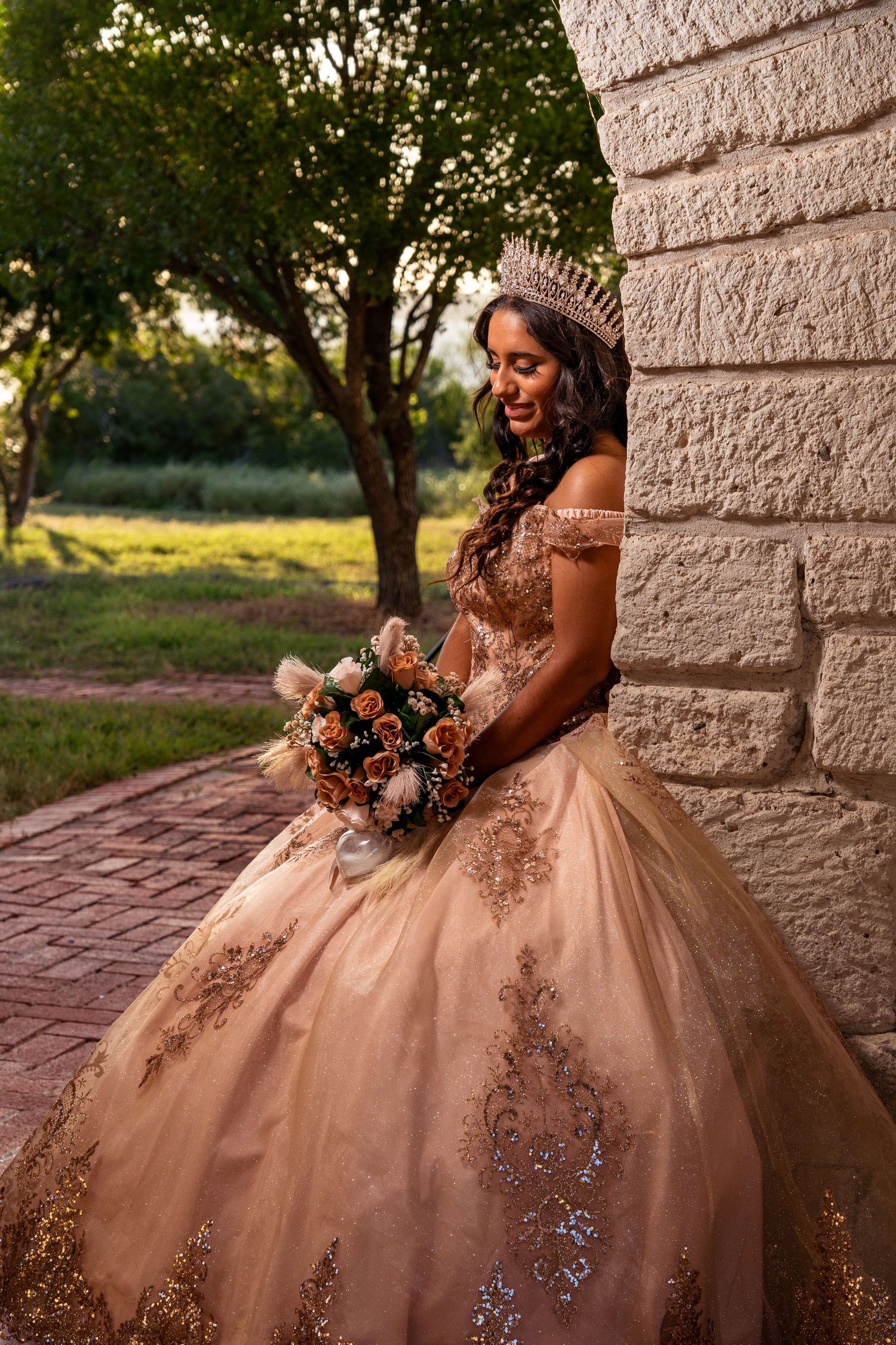 A quinceañera in a champagne ball gown with intricate beading poses beside a stone wall at golden hour.