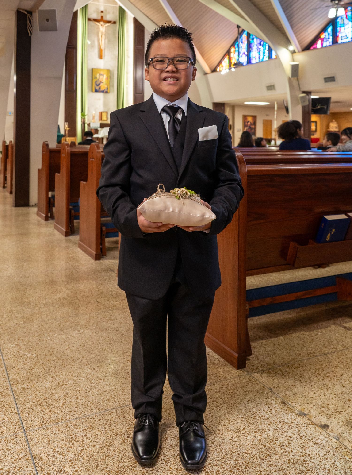 A person in a black suit stands in a church aisle holding a white pillow during a religious ceremony.