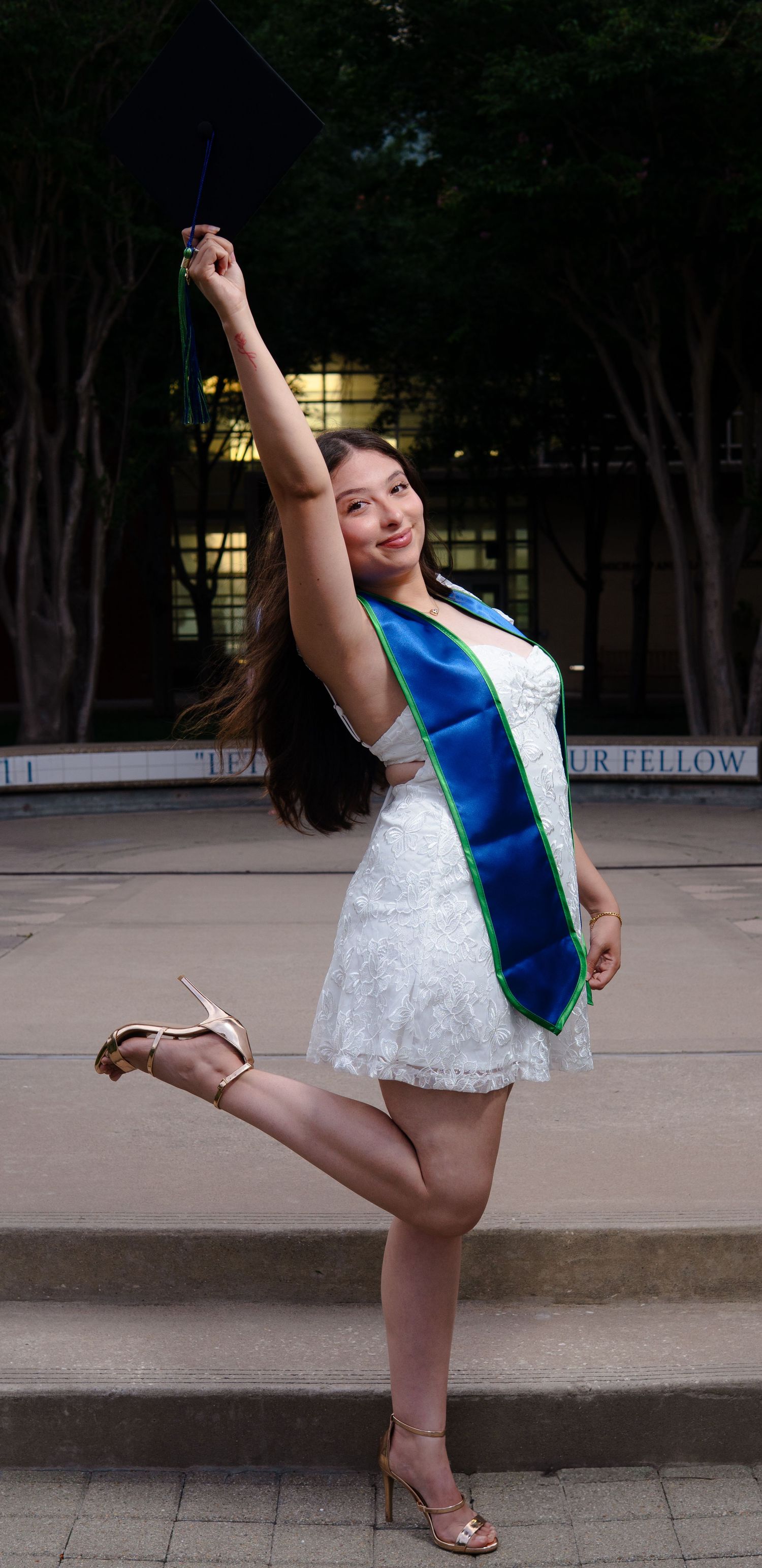 Graduate celebrating in white lace dress and blue sash strikes joyful pose on campus steps at dusk.