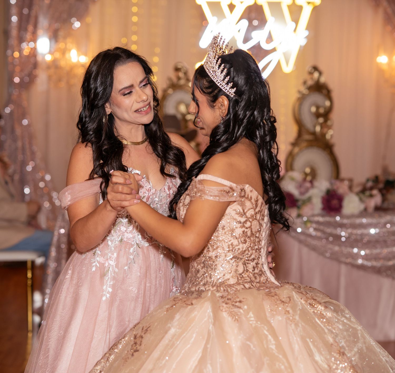 Two young women in elegant gowns share a dance at a quinceañera, one wearing a sparkling tiara and crown.
