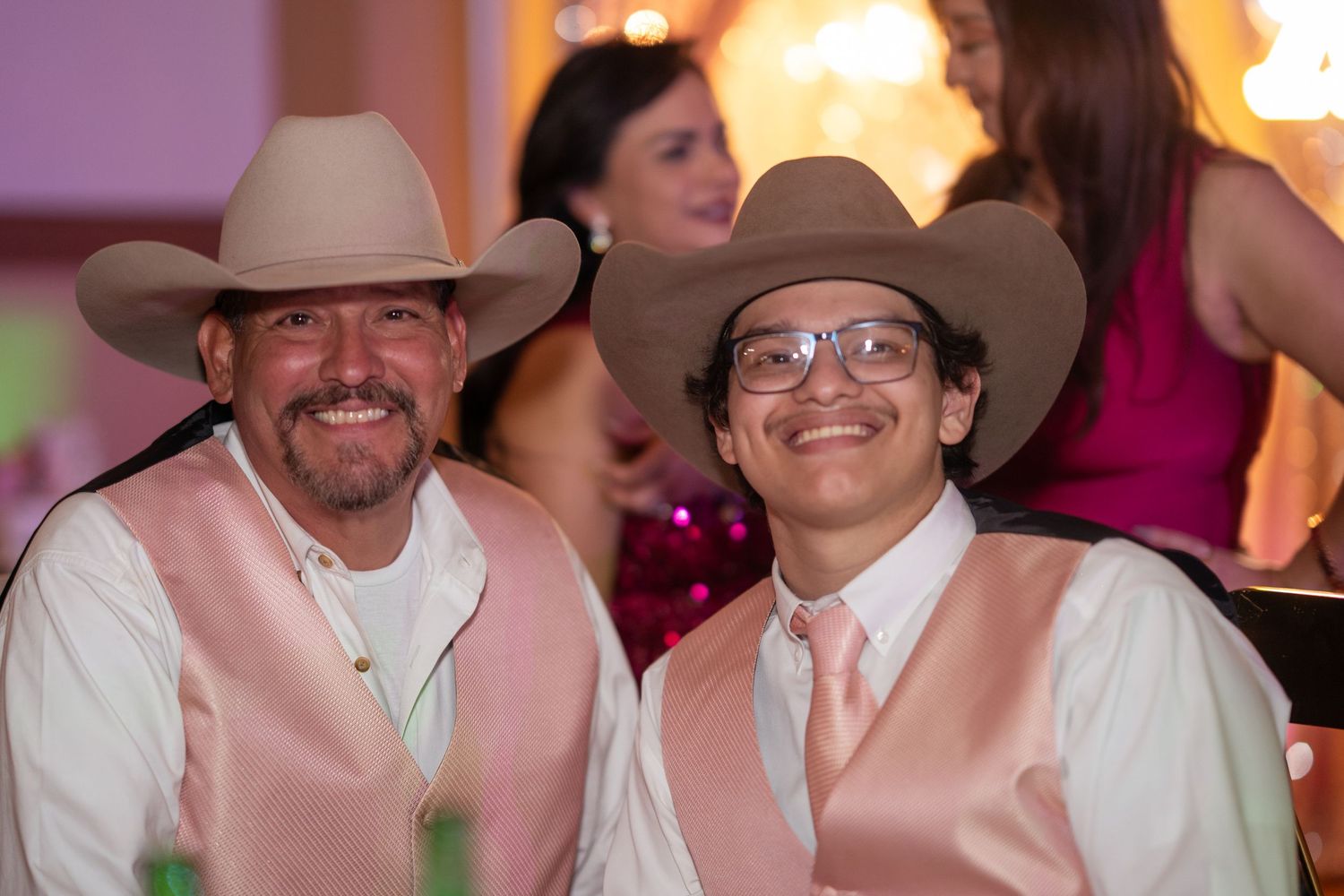 Two men in matching pink vests and cowboy hats smile together at a quinceañera celebration.