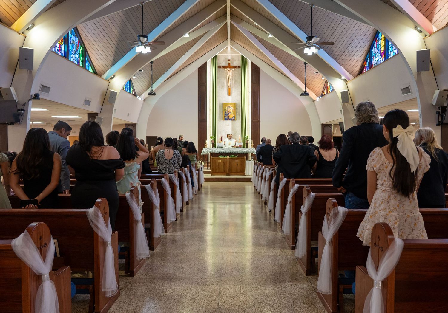Guests standing in church pews during a wedding ceremony in a beautiful Catholic church with stained glass windows.