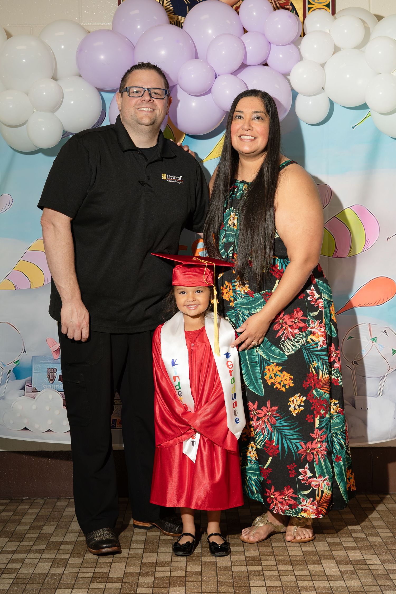 A kindergarten graduate in red cap and gown poses with parents in front of festive balloon backdrop.