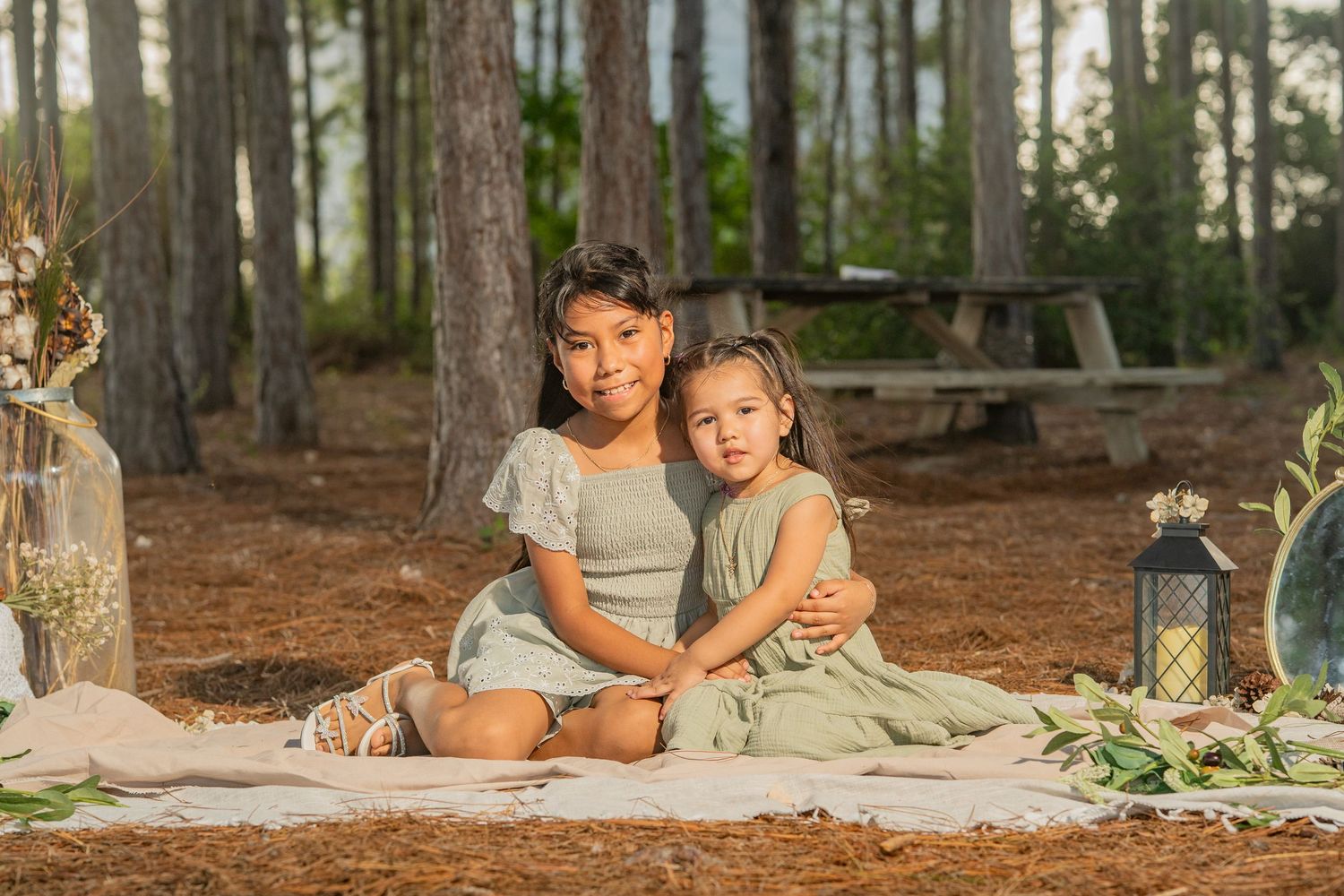 Two young sisters in sage green dresses sit together on a blanket in a pine forest during a styled outdoor photo session.