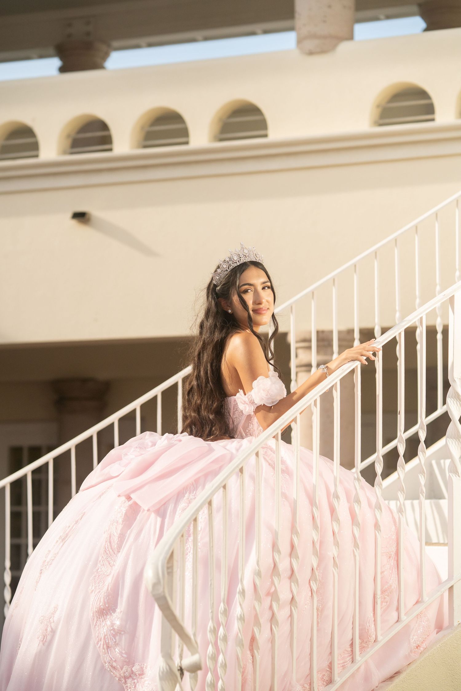 Young woman in pink quinceañera ball gown and tiara poses gracefully on white staircase outdoors.