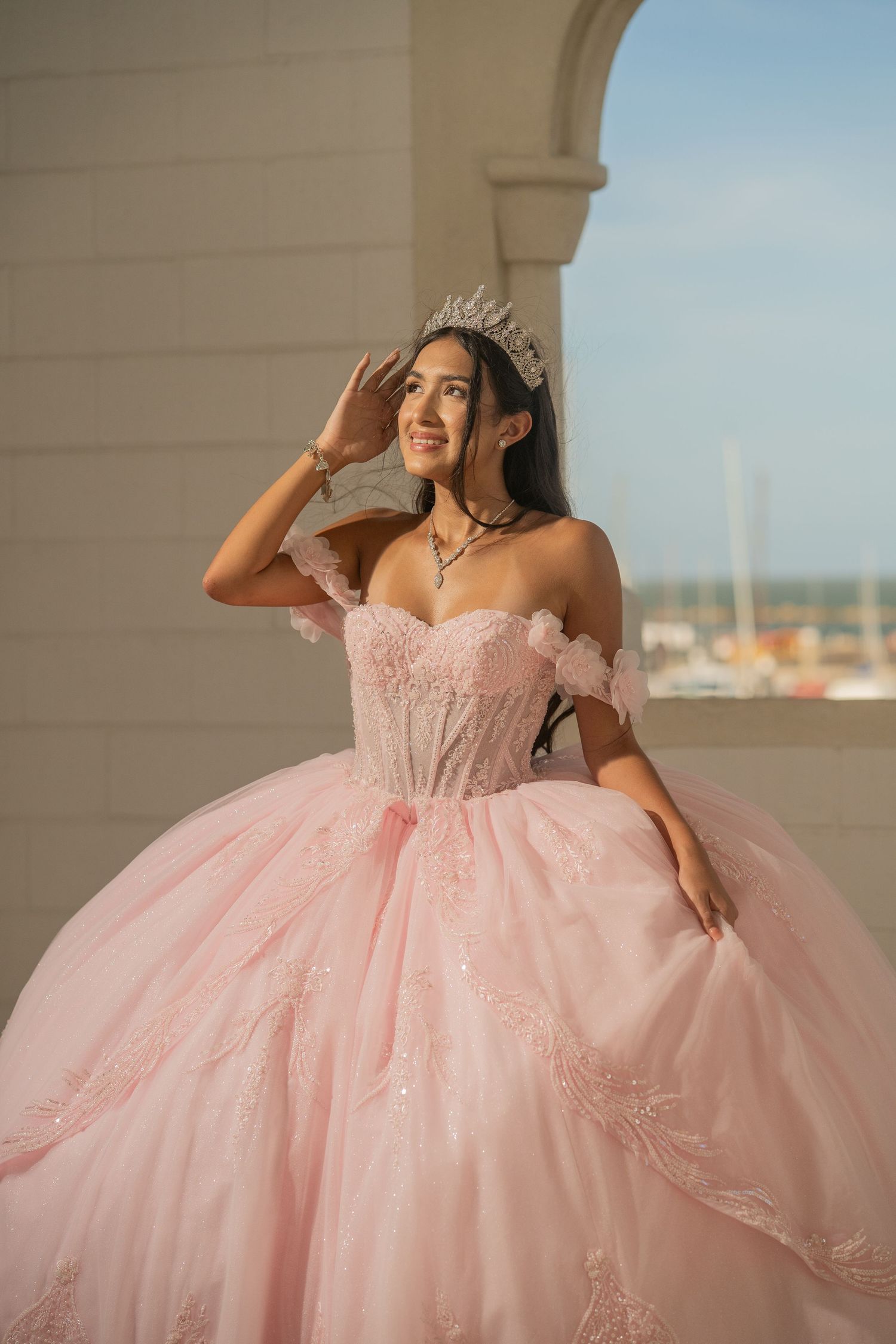 Smiling young woman in elegant pink quinceañera dress and silver tiara posing outdoors on a balcony.