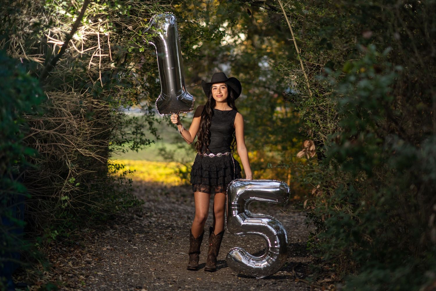 Teen girl in black dress and cowboy hat holds silver number 15 balloons on a forest path for quinceañera photos.