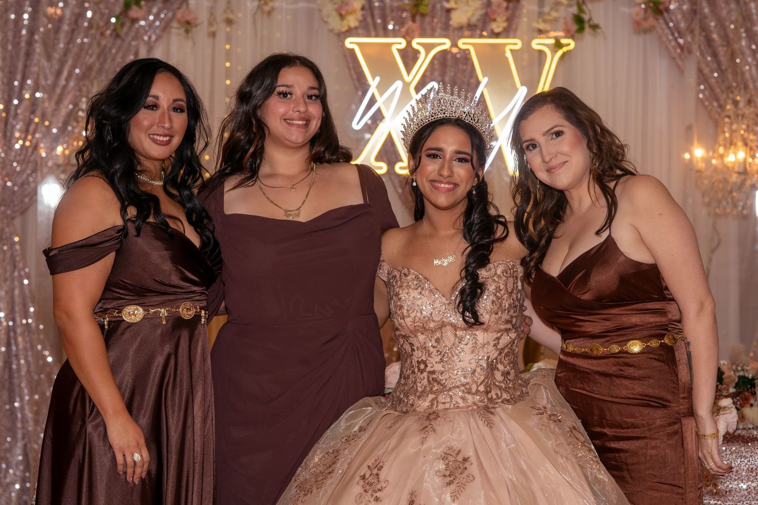 Four women pose together at a quinceañera celebration with XV backdrop and sparkling decorations.