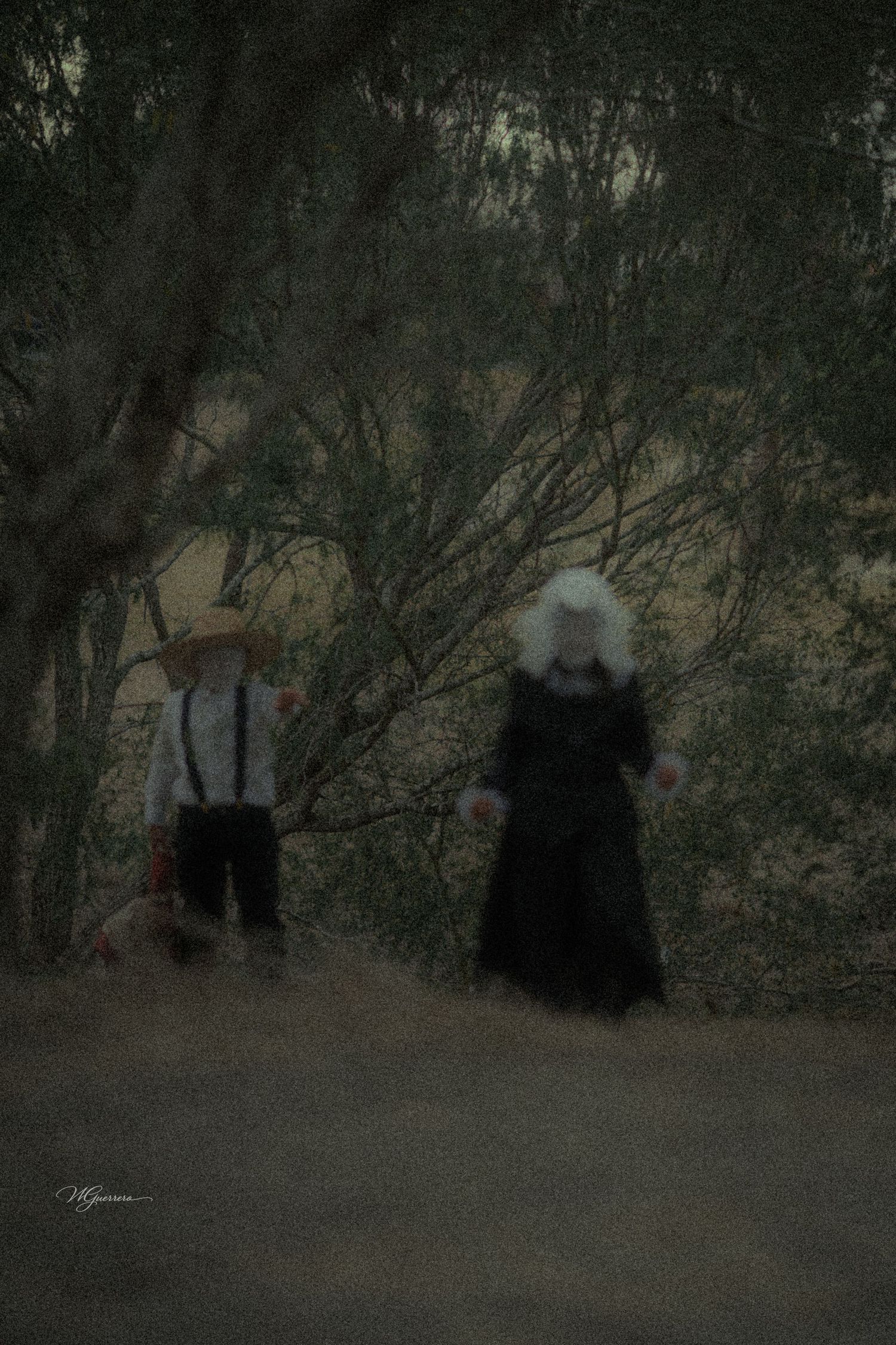 A boy in suspenders and straw hat walks with a woman in a long black dress through a dark wooded area.