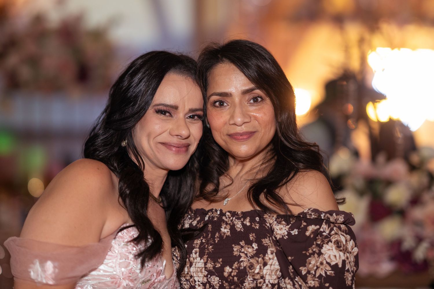 Two smiling women pose cheek to cheek at an elegant evening celebration with warm bokeh lighting behind them.