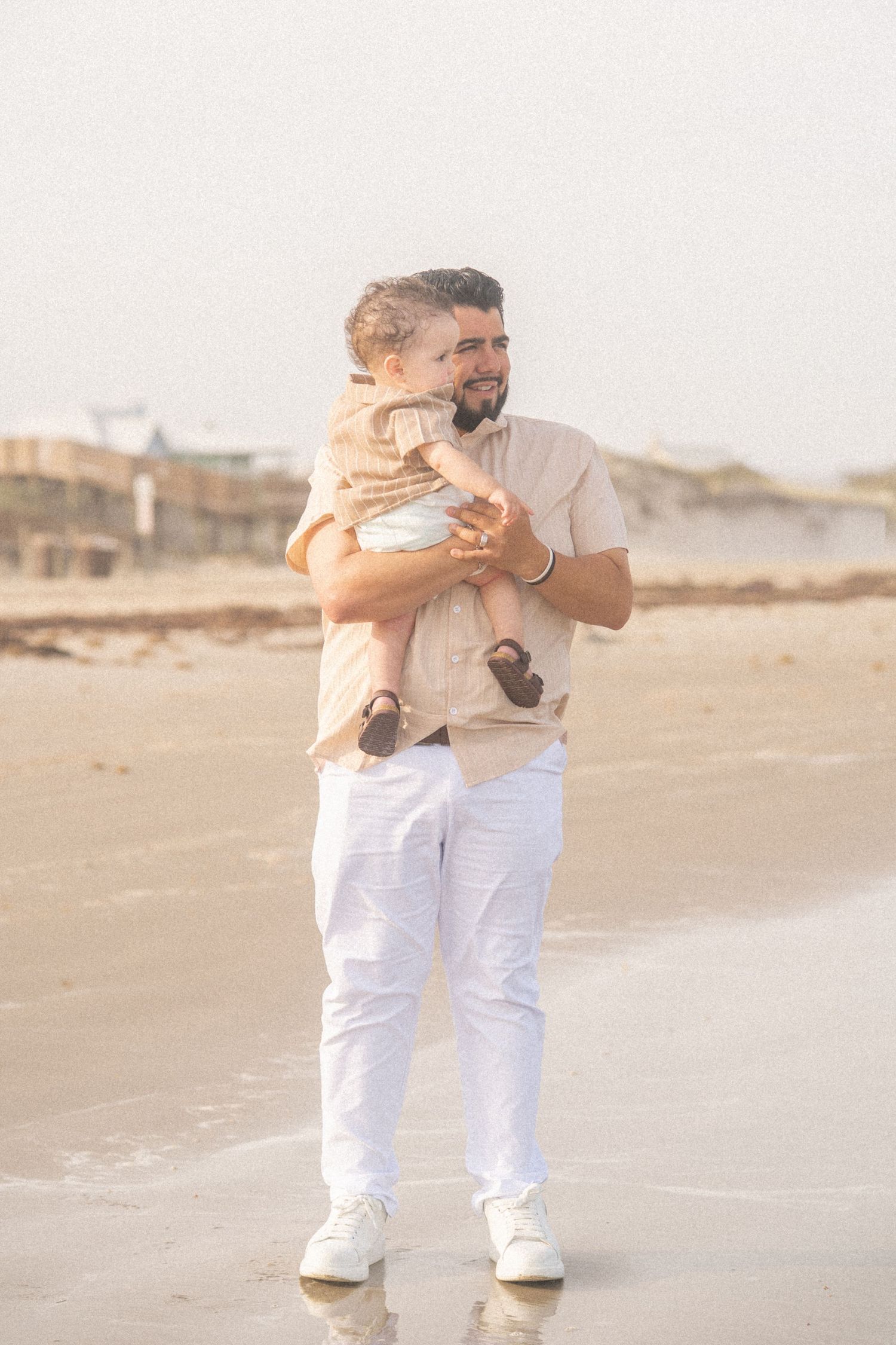 A bearded father holds his baby on a sunny beach, both dressed in neutral tones with white pants.