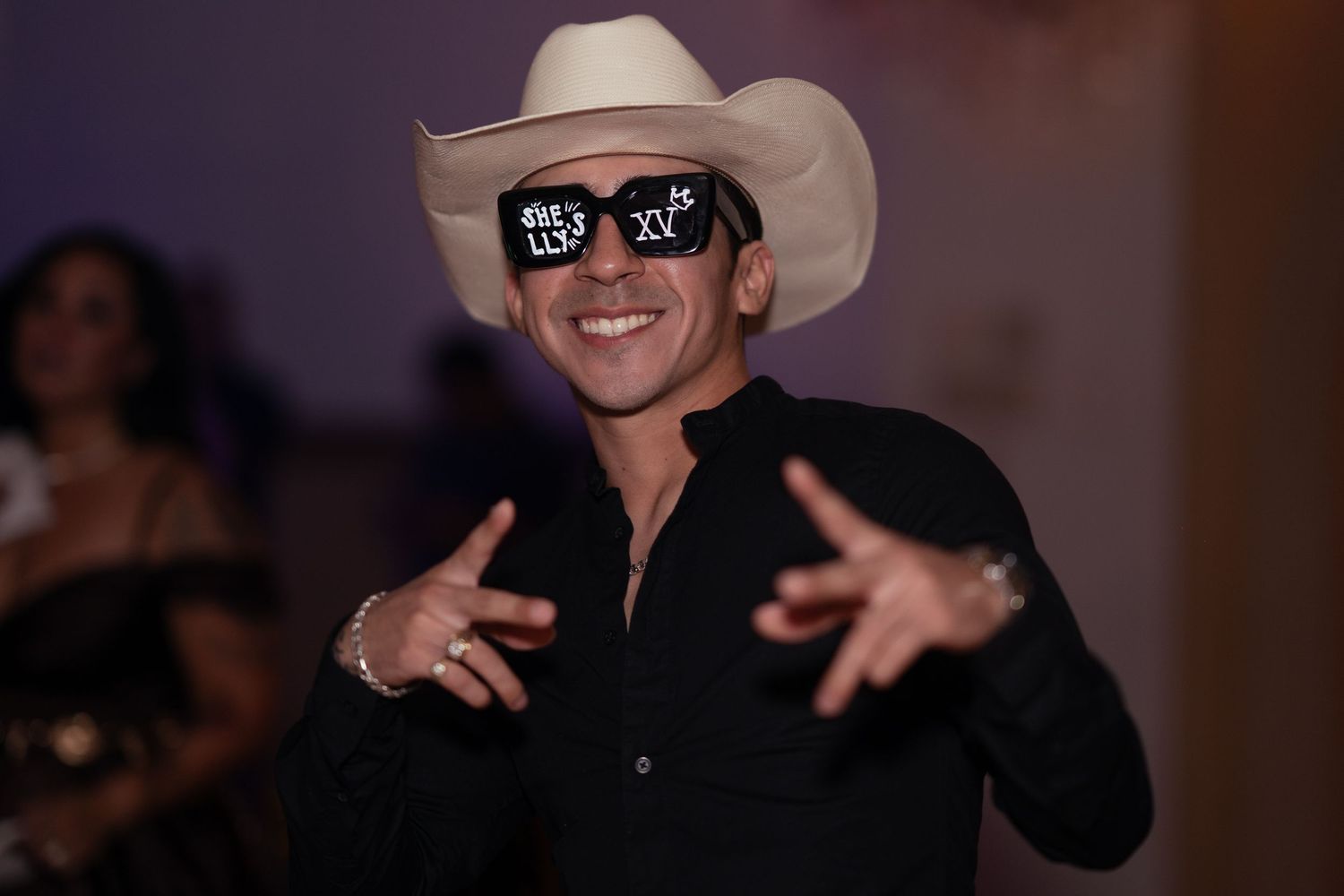 Man in white cowboy hat and black shirt wearing XV años novelty sunglasses celebrates at a quinceañera party.