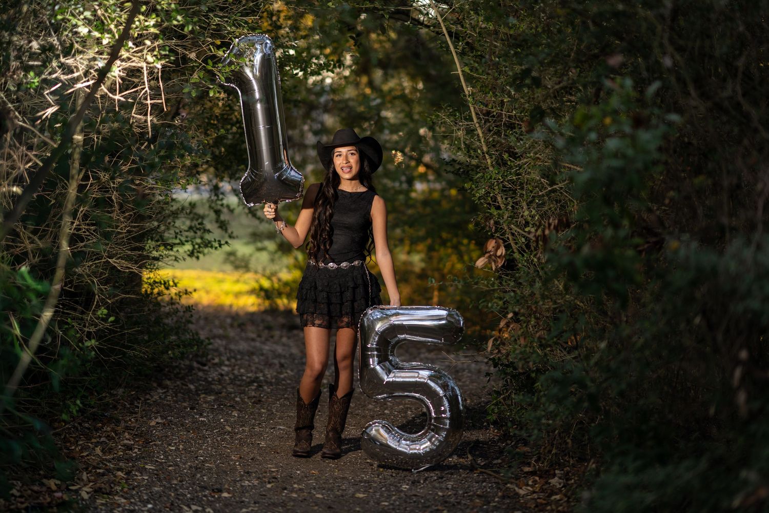 Teen girl in black dress and cowboy hat holds silver number 15 balloons on a forest path.