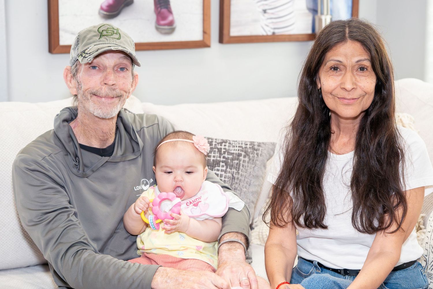 A man and woman sitting on a couch smiling, holding a baby girl in a bright living room.