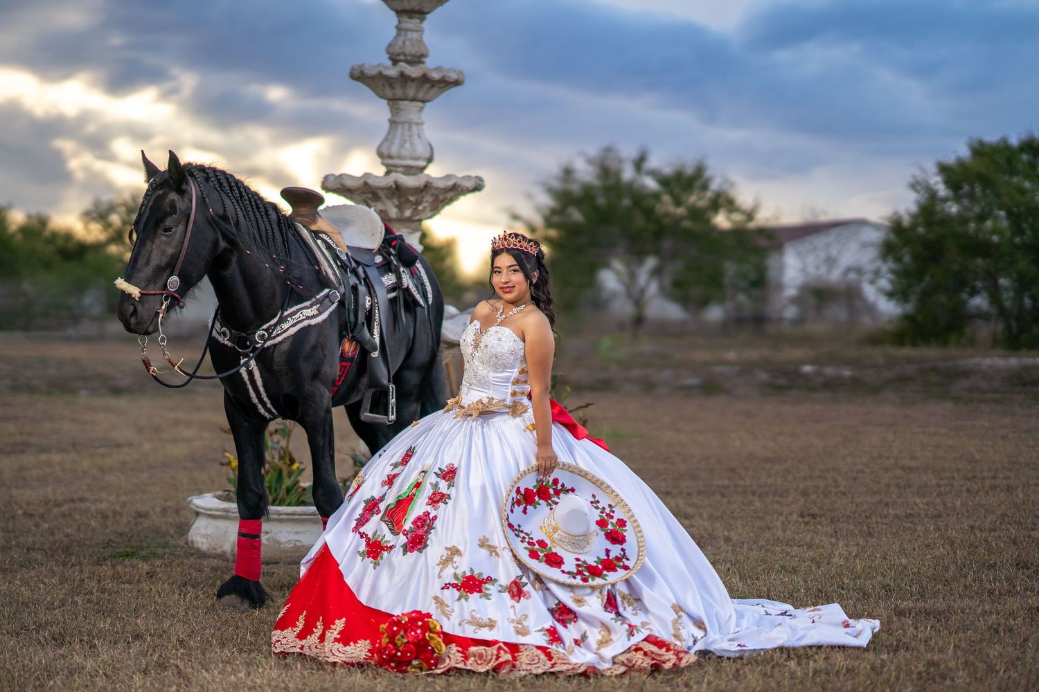White and red embroidered quinceañera gown spread out on grass with dramatic cloudy sky overhead.