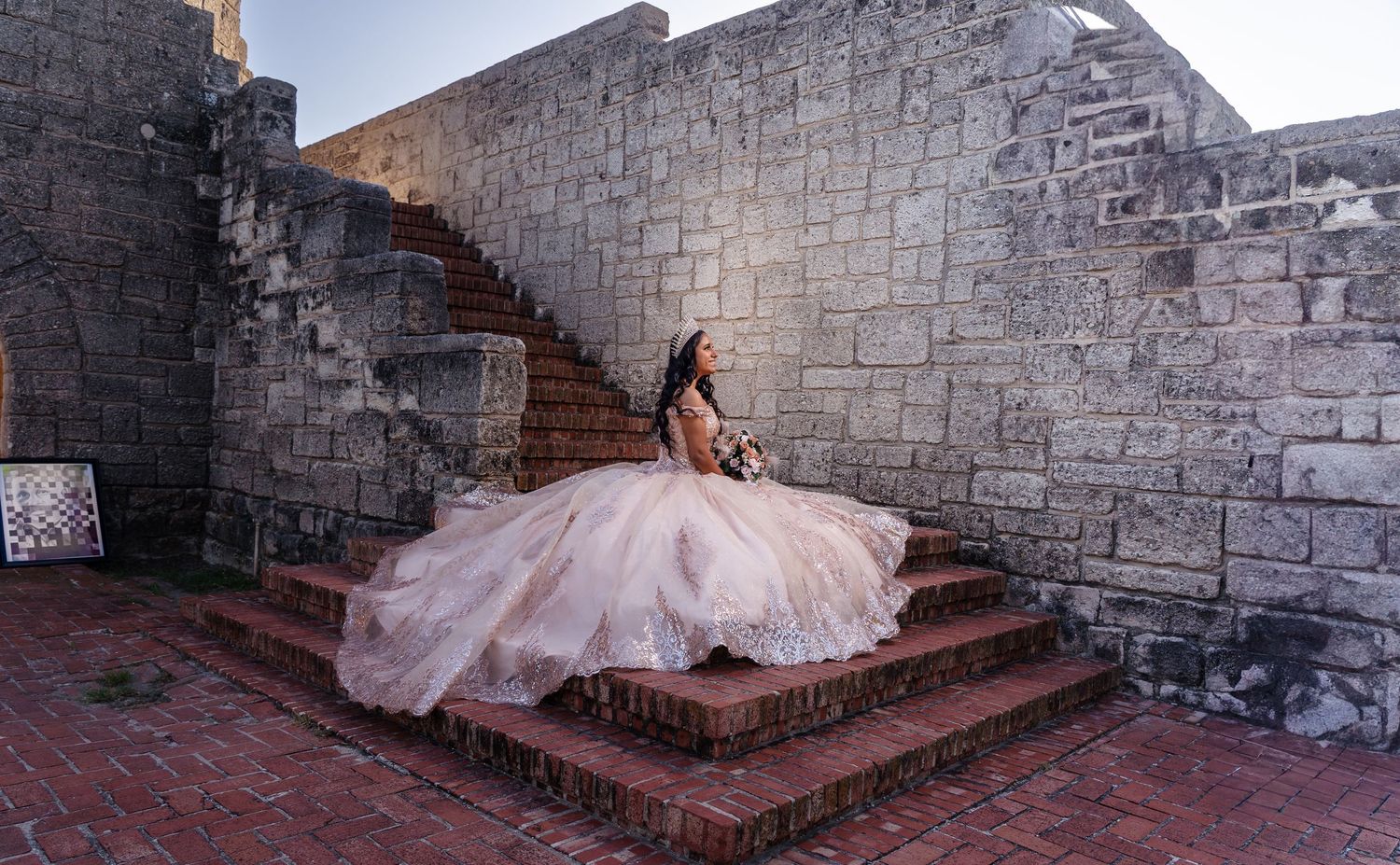 A stunning blush pink quinceanera dress with intricate beading photographed on stone stairs against a historic wall.