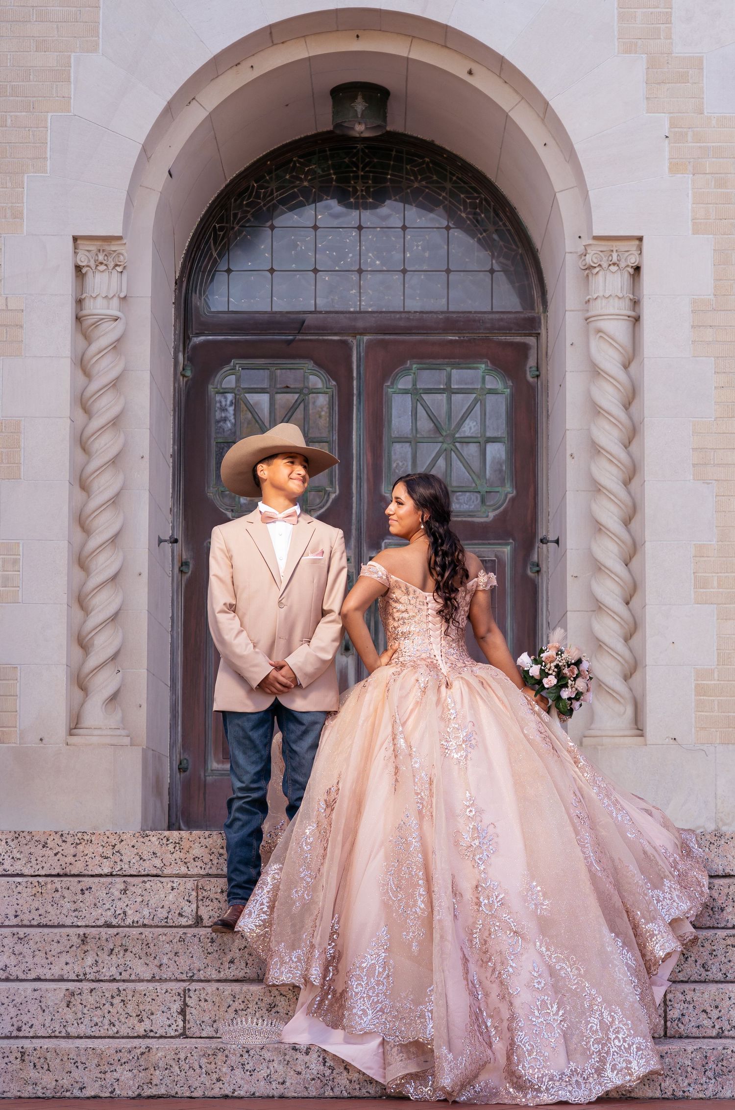 A couple in formal attire poses on stone steps in front of an ornate archway, with the woman wearing a blush ballgown.