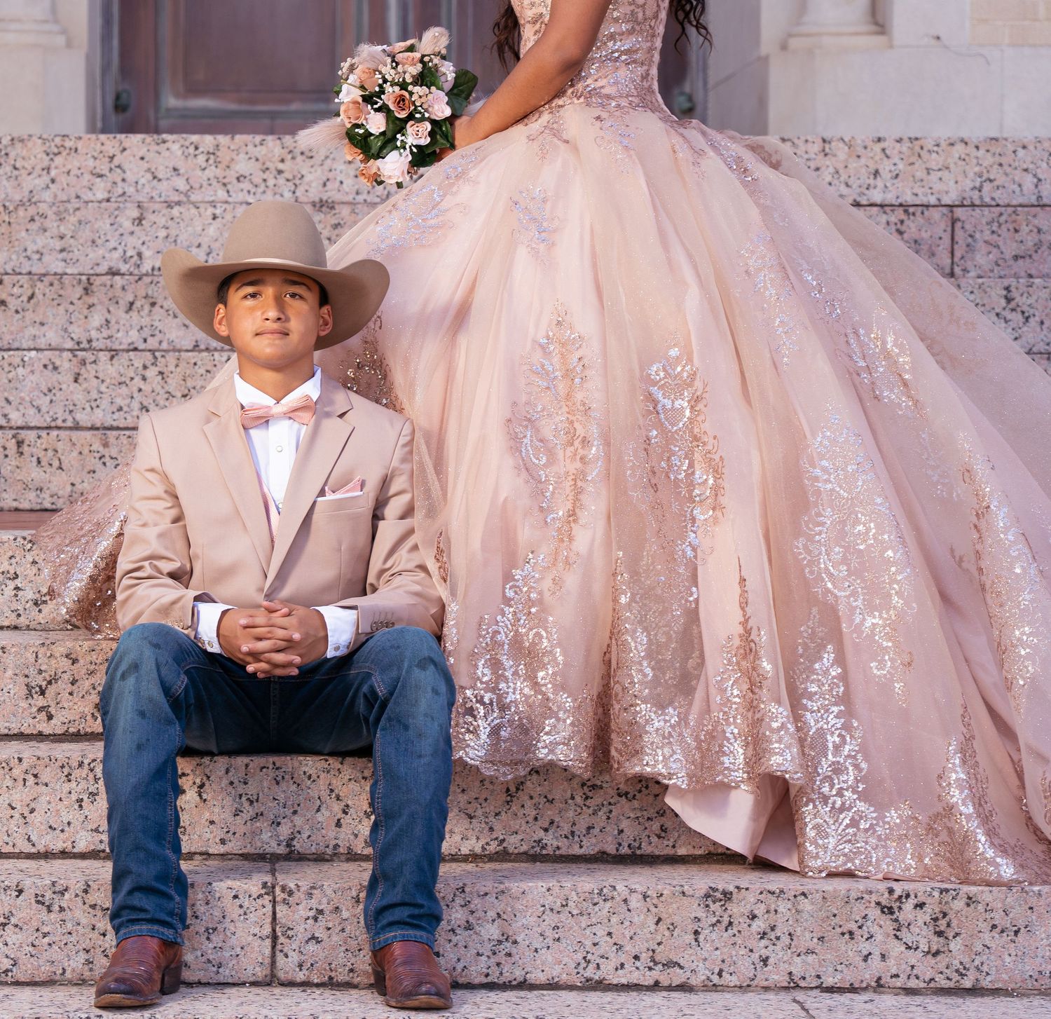 A young boy in a cowboy hat sits next to a flowing pink lace dress on stone steps at sunset.
