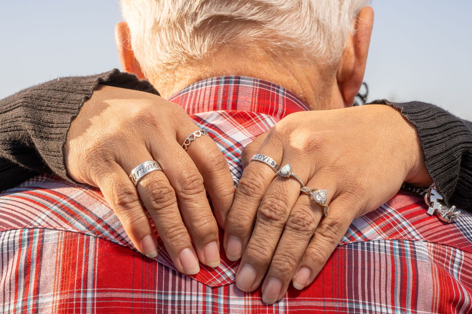 Elderly couple embracing, showing hands with silver rings on a red plaid shirt.