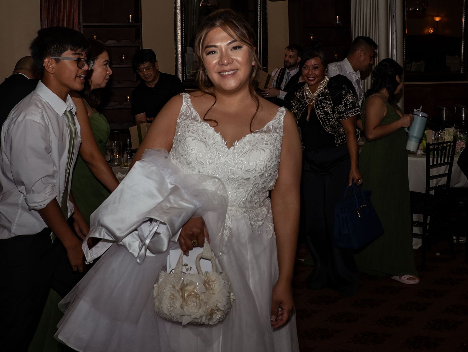 A young woman smiles in a white lace quinceañera gown holding a floral purse at a celebration.