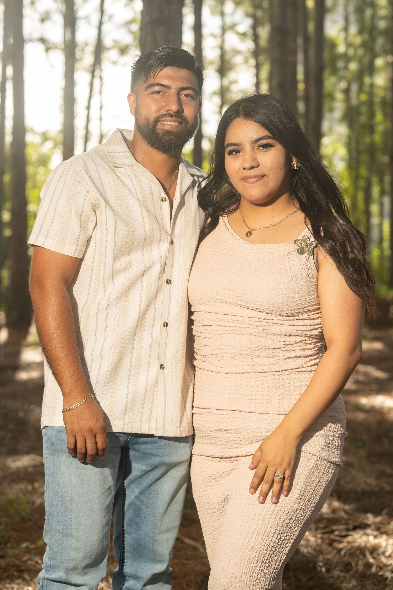 A smiling couple poses together in a sunlit forest, man in striped shirt and woman in pink dress.