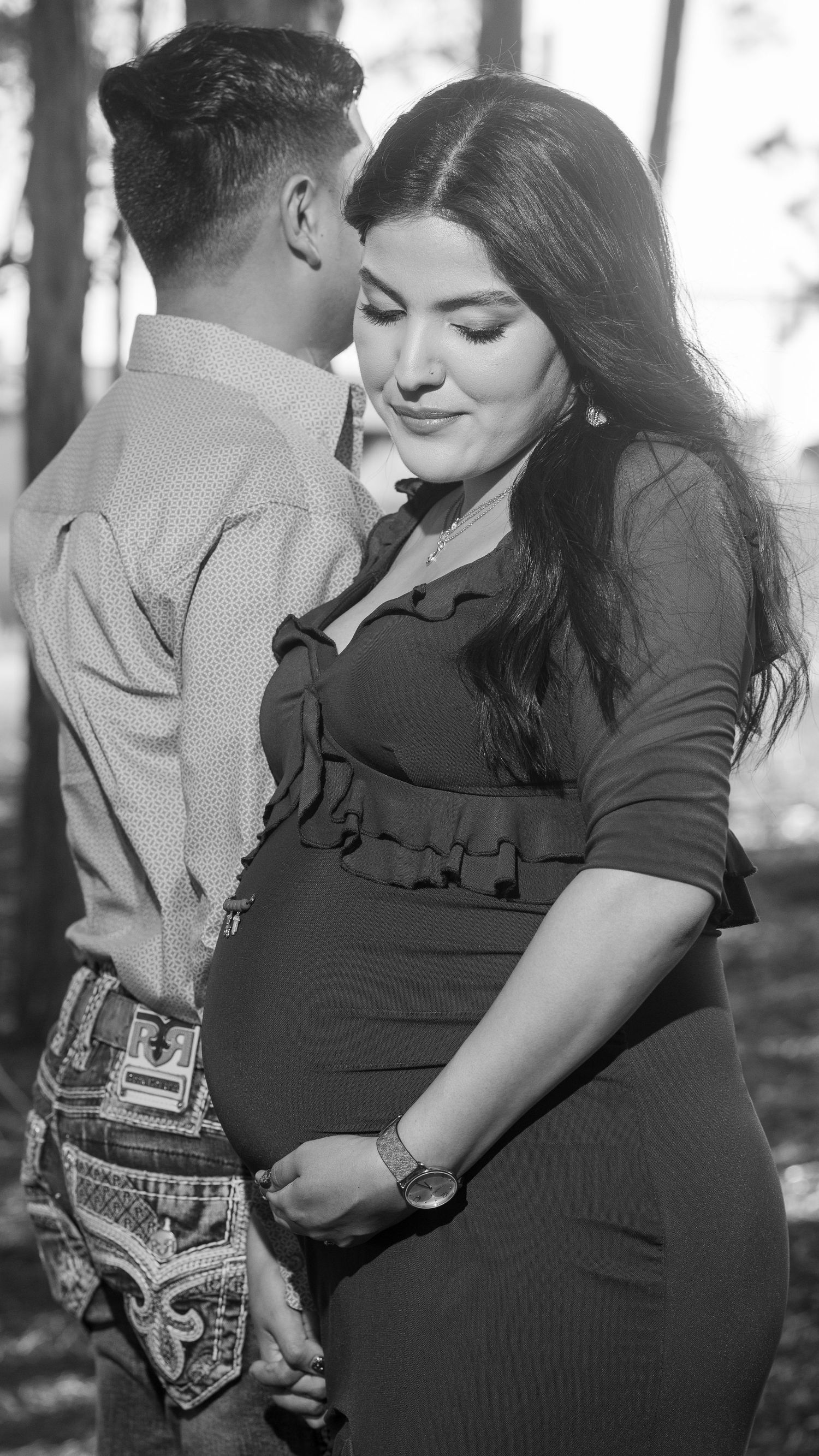 Black and white maternity photo of a pregnant woman in a fitted dress holding hands with her partner outdoors.