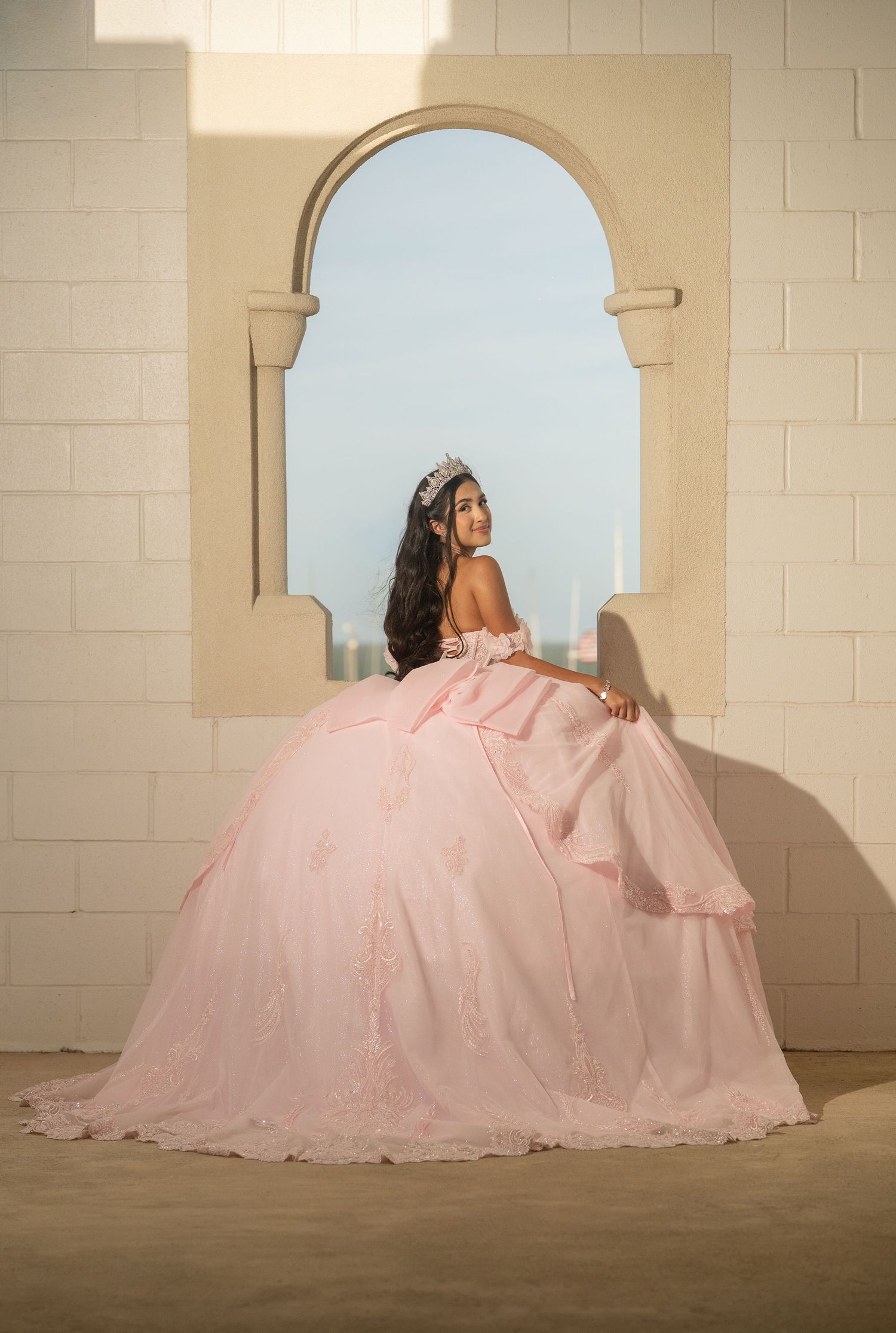 Young woman in a pink quinceañera ball gown and tiara posing by an arched window in golden light.