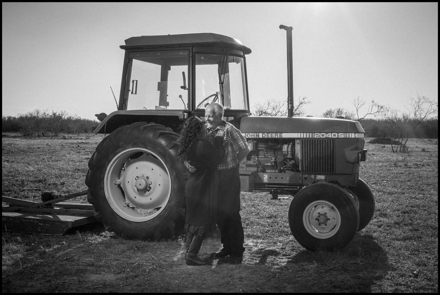 A farmer stands proudly beside a John Deere tractor in a rural field, captured in black and white photography.