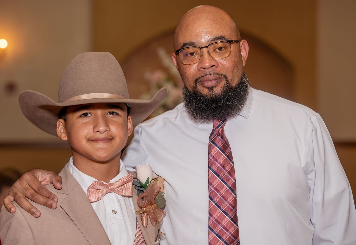A bald man with a beard and glasses poses with a boy wearing a cowboy hat and pink bow tie at a formal event.