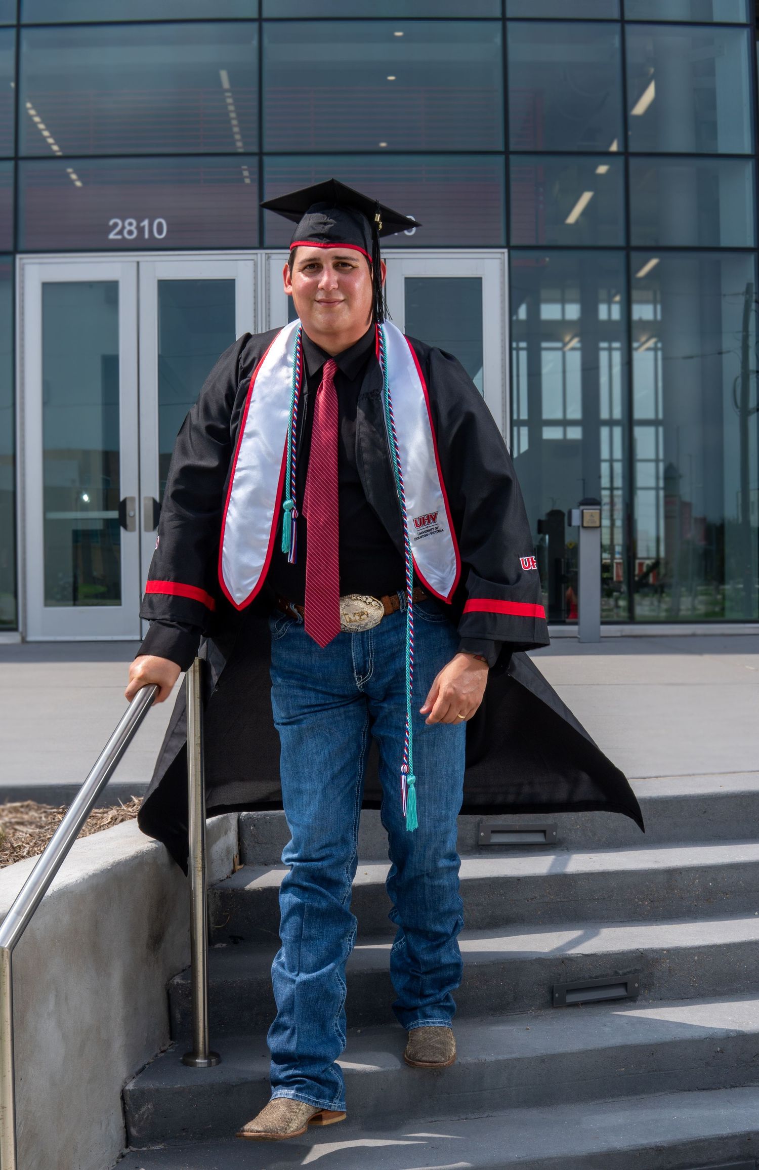 A graduate in cap and gown poses on steps outside a modern building with glass doors and walls.