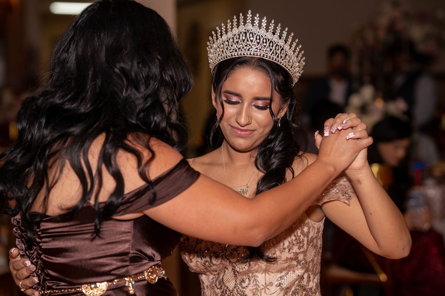 Young quinceañera in gold gown and crystal crown shares a dance with a guest at her celebration.