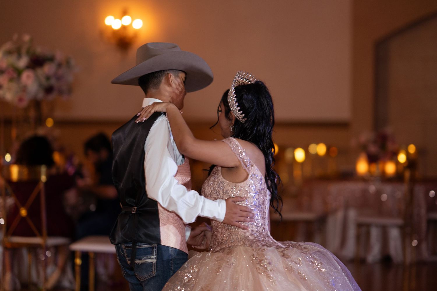 A young woman in a rose gold quinceañera gown and tiara shares a dance with a man in a cowboy hat.