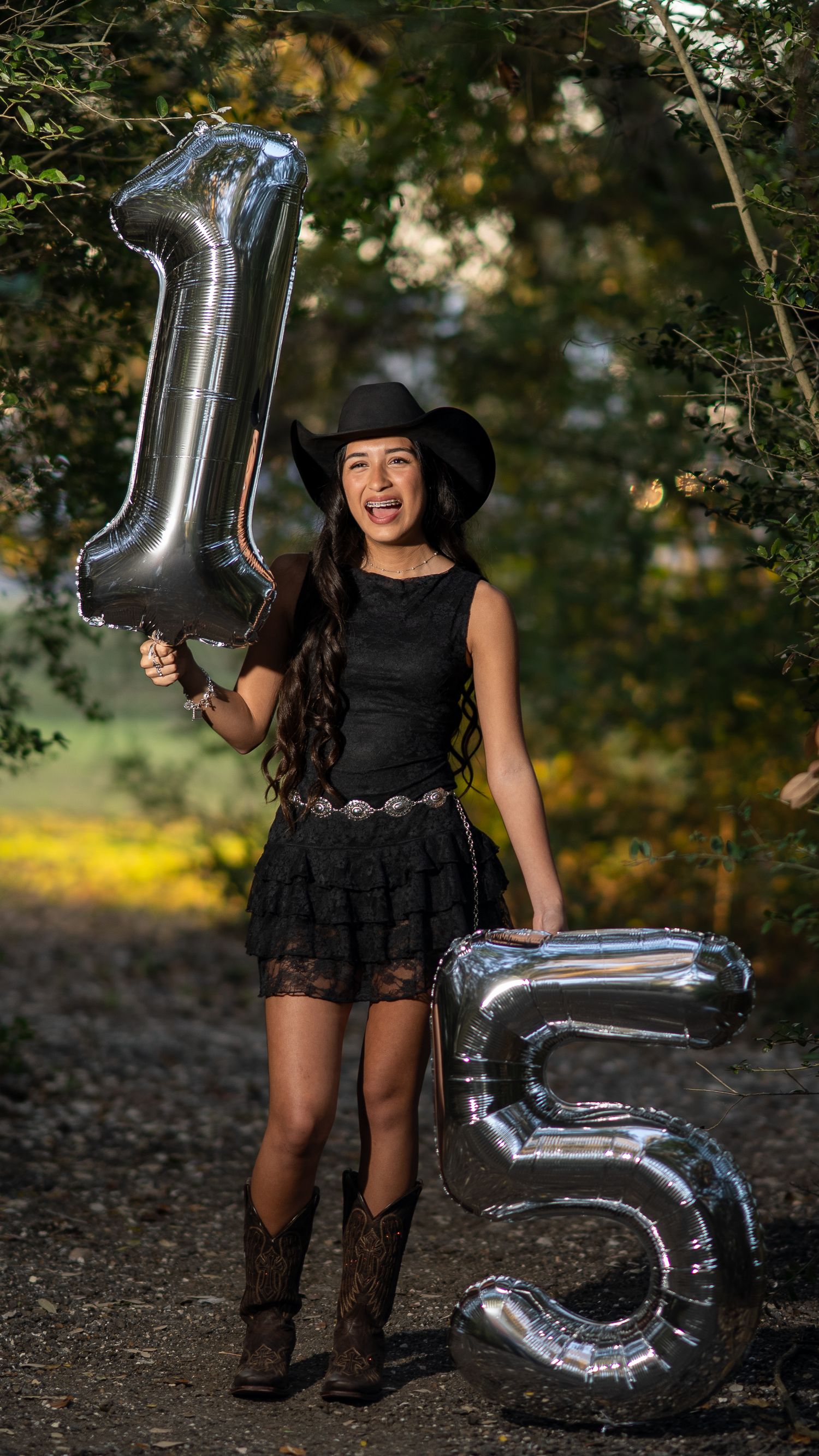A teenage girl in a black dress and cowboy hat holds silver number 15 balloons celebrating her Quinceañera outdoors.