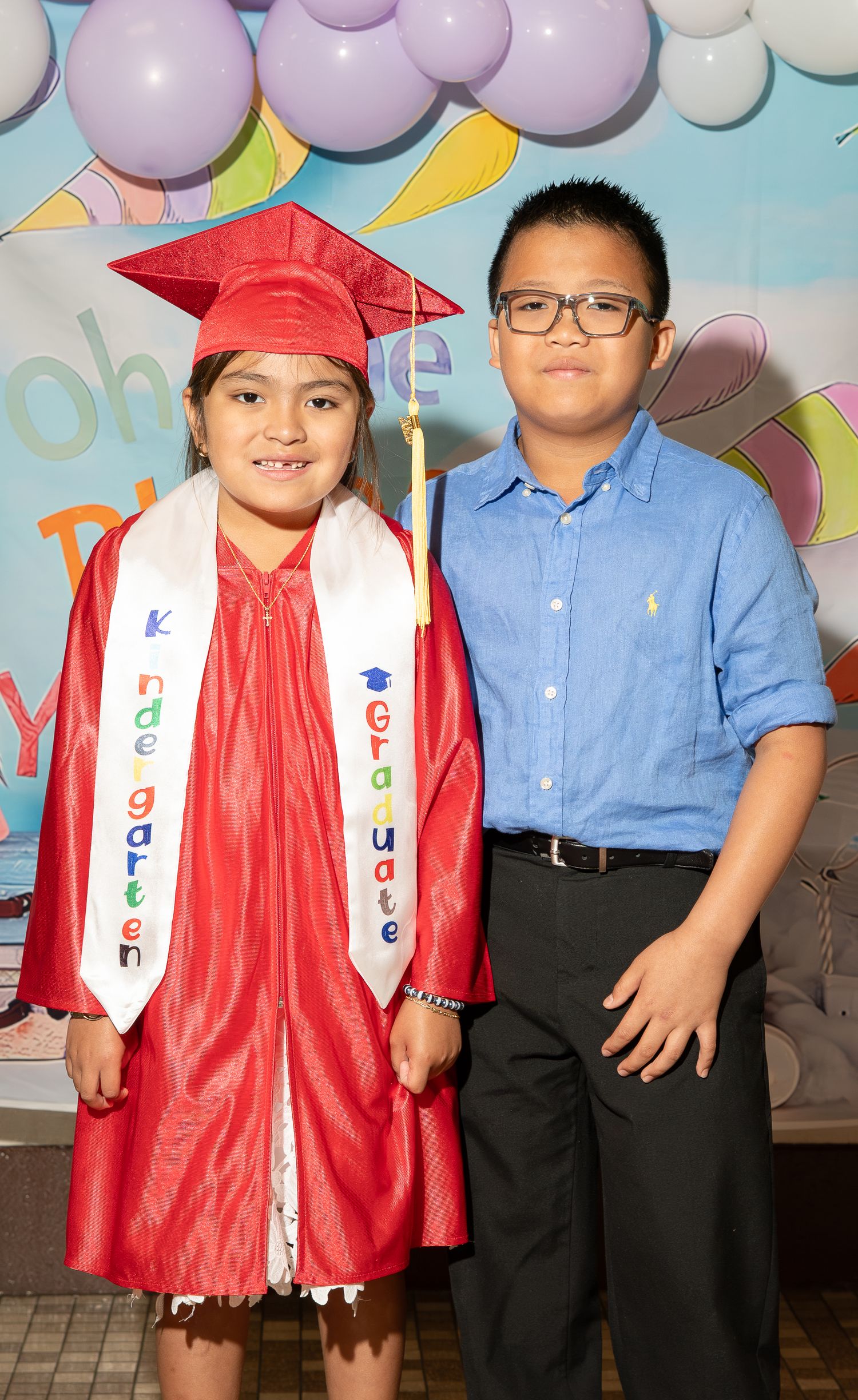 Young girl in red kindergarten graduation cap and gown poses with older boy in blue shirt at graduation ceremony.