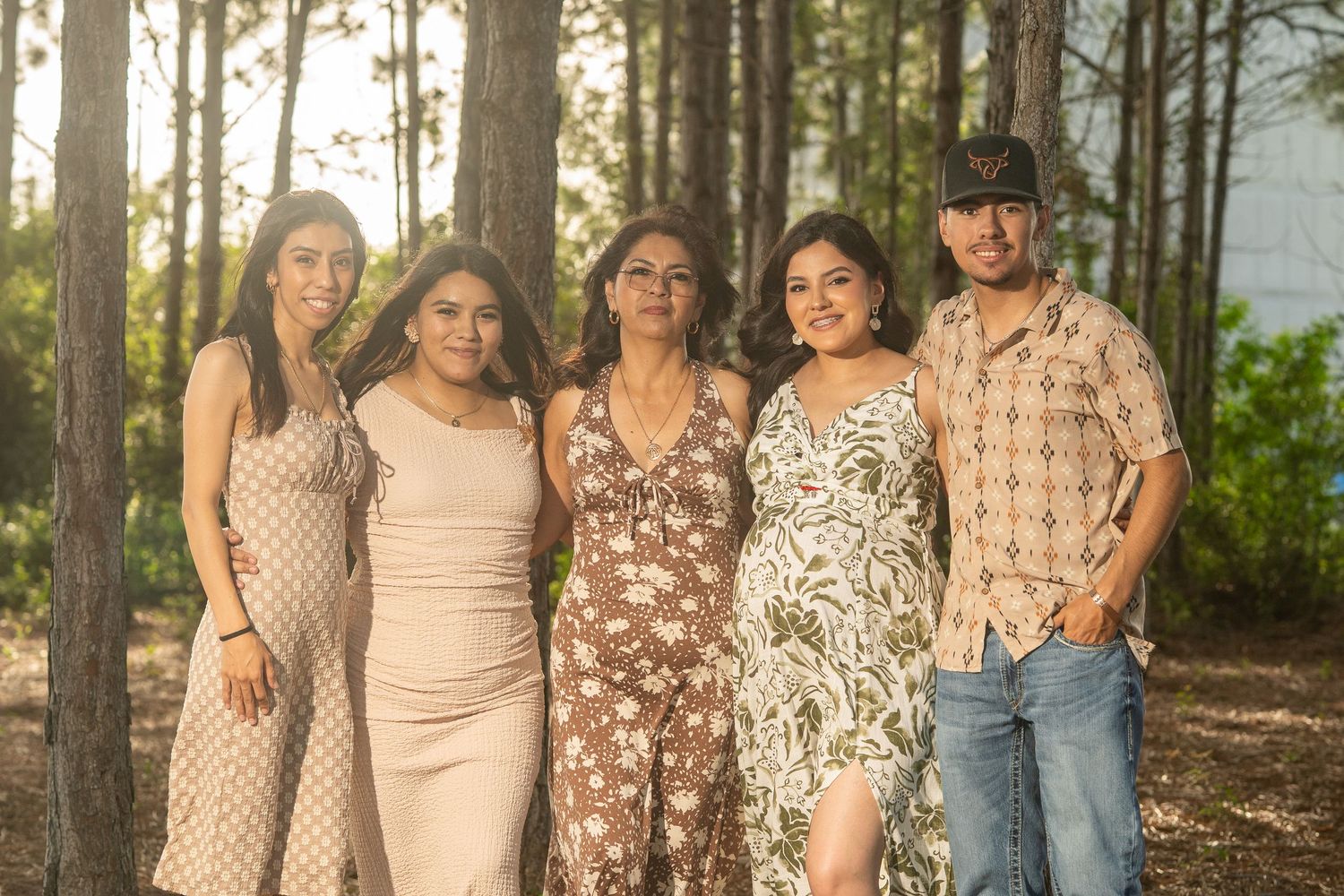 Five people posing together outdoors among tall pine trees in warm golden light, dressed in casual summer attire.