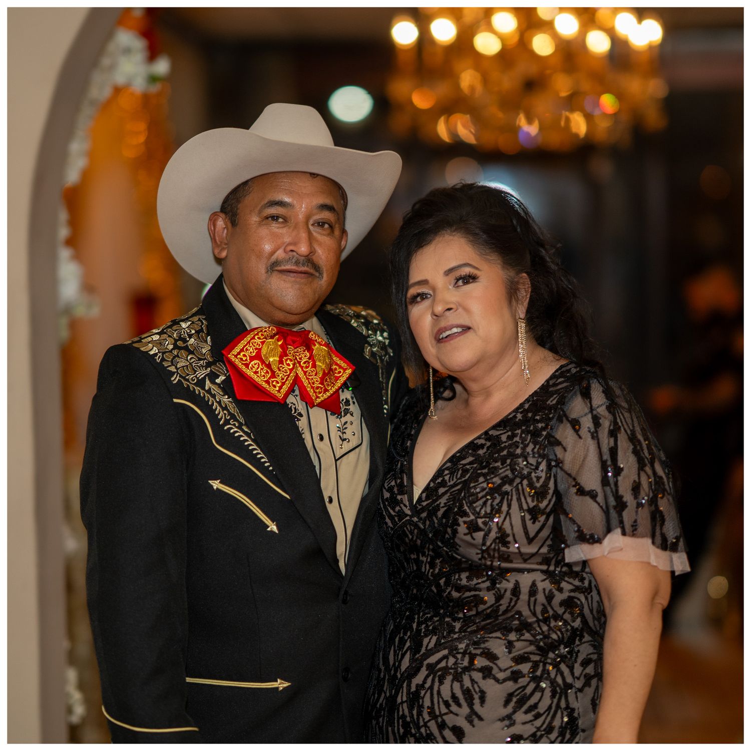 A couple in western attire, with a person wearing a cowboy hat and embroidered jacket posing with someone in a black dress.
